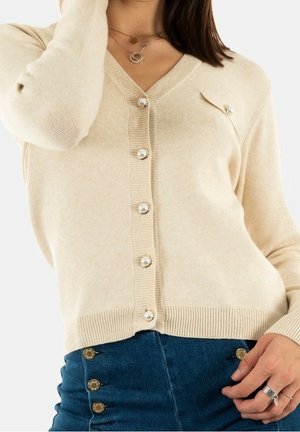 Frau trägt eine beige Strickjacke mit perlenähnlichen Knöpfen und einen blaue, hoch taillierte Rock mit goldenen Knöpfen, ergänzt durch Ringe und mehrschichtige Halsketten.