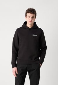 Czarny hoodie z miękkiego materiału, wyposażony w przednią kieszeń, ściągacze przy mankietach oraz małe szare logo Calvin Klein na klatce piersiowej.