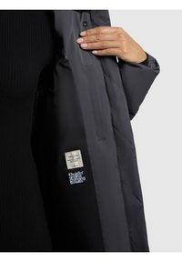 Vycpaná černá bunda s hladkým, lesklým materiálem. Viditelné štítky uvnitř uvádějí "Vyrobeno s láskou" a "Khujo Original Changing Threads."