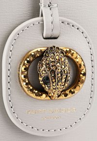 Etichetta in pelle di grigio pallido con hardware dorato, arricchita da embellishments testurizzati e un centro in pietra nera. "Kurt Geiger London" impresso in basso.