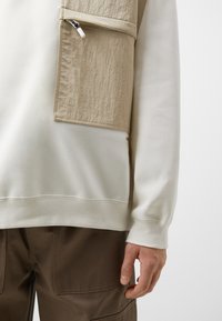 Persona con sudadera color crema con bolsillo en el pecho beige y pantalones marrones, con la mano relajada a un lado sobre un fondo liso.