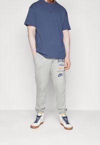 Marinblå t-shirt med en liten logotyp, grå sweatpants med tryckta detaljer, och vita högstövlade sneakers med blå snörning och bruna sulor.