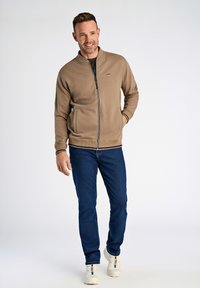 Giacca beige con zip e colletto alto, due tasche laterali e polsini a coste, abbinata a jeans blu scuro e sneakers bianche.