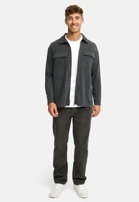 Grey corduroy shirt jacket with two front pockets, worn over a white crew-neck shirt, paired with dark pants and white sneakers.