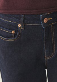 Tmavě modré džíny z denimu s žlutým prošitím, měděnými knoflíky a klasickým designem s pěti kapsami. Hladká textura s přiléhavým střihem.