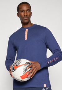 Topp navy med lange ermer og rosa detaljer ved halslinning og ermer, med teksten "NIKE" og "BARÇA"; holder en mønstret fotball.