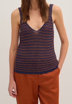 Frau trägt ein ärmelloses, marineblau-oranges gestreiftes Strick-Top mit rostfarbenen Hosen und steht mit einer Hand in der Tasche.
