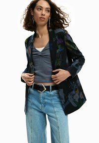 Blazer nero con stampa floreale, top grigio a coste, jeans azzurri a vita alta e cintura nera con fibbia geometrica argentata.