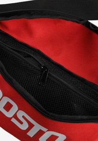 Czerwona fabriczna torba z czarnym zamkiem błyskawicznym i siateczkową kieszenią. Wnętrze jest czerwone o gładkiej fakturze. Na zewnątrz widoczne jest wyraźne białe logo.