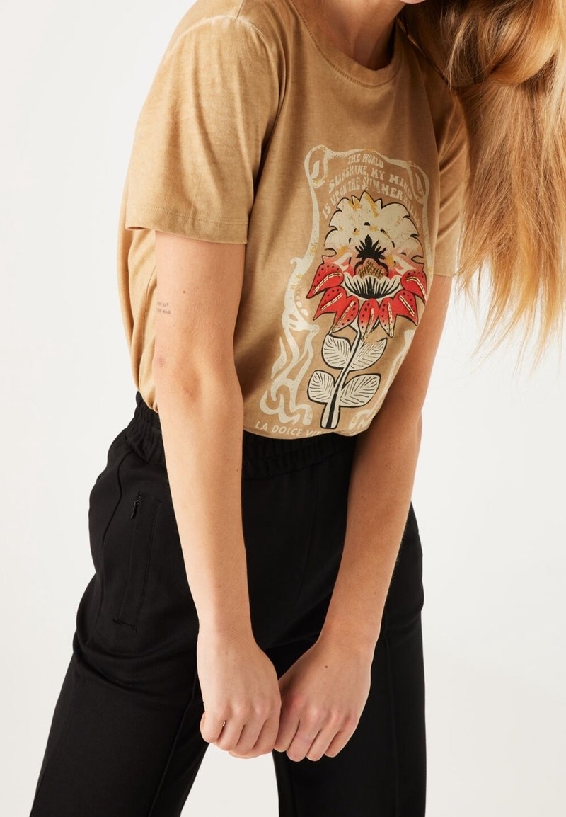 Camiseta de algodón marrón con un gráfico floral que presenta detalles en rojo y negro, combinada con pantalones negros de cintura elástica con bolsillo lateral.