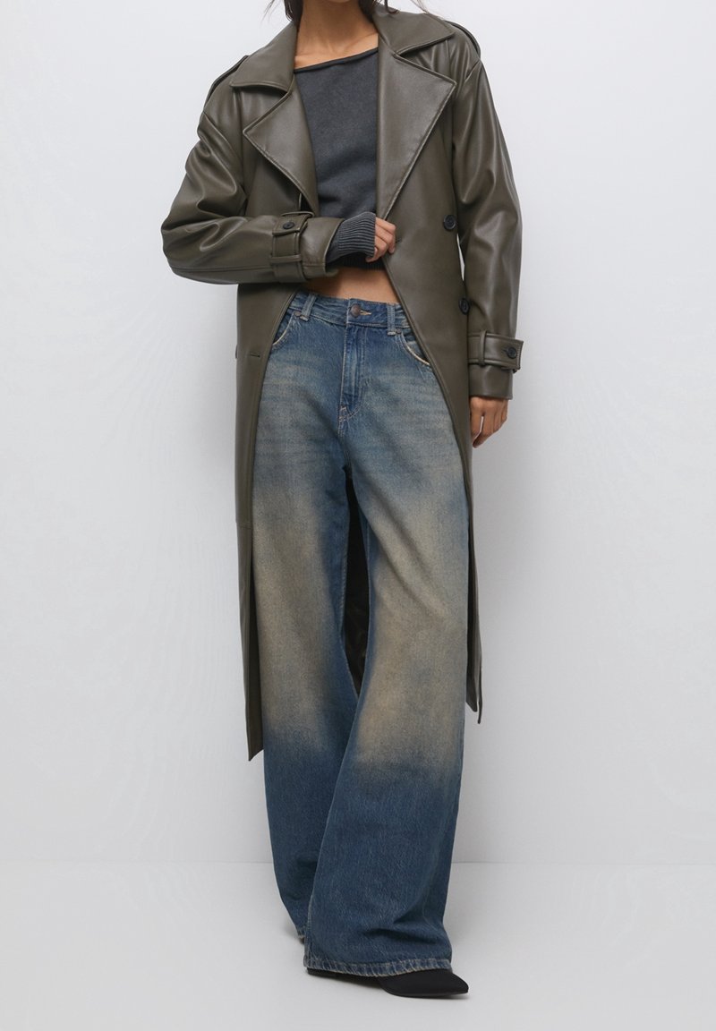 Personne portant un long manteau en cuir vert foncé, un haut court foncé et un jean bleu délavé à jambes larges, debout devant un fond clair uni.