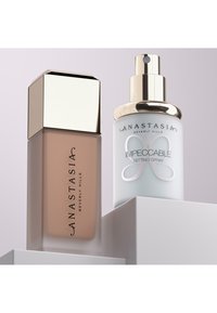 Twee cosmetische producten: een rechthoekige glazen foundationfles met een gouden dop en een ronde setting sprayfles met een zilveren logo en pomp.