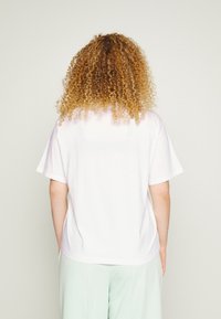 Camiseta blanca de algodón con un corte holgado, mangas cortas y un pequeño logo en la espalda. Combinada con pantalones verdes claros.