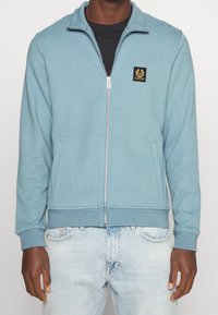 Sudadera con cremallera en azul claro, mezcla de algodón; cuenta con bolsillos laterales y puños de canalé. Parche del logo negro en el lado izquierdo del pecho. Ajuste casual.