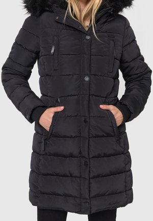 Femme portant un manteau d'hiver matelassé noir avec poches avant et une capuche bordée de fourrure noire, mains dans les poches.