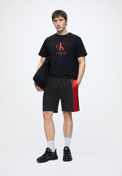 Млад мъж стои, облечен в черна тениска Calvin Klein, черни къси панталони с червена ивица, бели чорапи и черни маратонки, държейки черно яке.