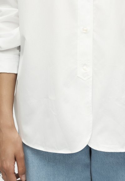 Άτομο που φοράει άσπρο πουκάμισο με κουμπιά και τυλιγμένα μανίκια καθώς και μπλε τζιν, δείχνοντας την περιοχή της μέσης και το αριστερό χέρι.