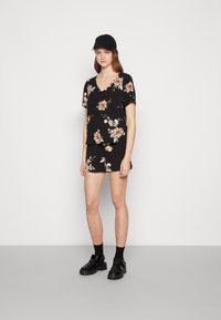 Top preto de flores com mangas curtas e decote em V, combinado com calções a condizer. Usado com calçado preto e meias, contra um fundo neutro.
