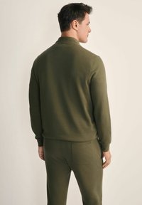 Mężczyzna odwrócony plecami, ubrany w dopasowany oliwkowozielony długi sweter i spodnie, stojący na tle jasnej, gładkiej ściany.