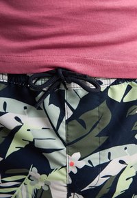 Camicetta rosa a maniche corte sopra pantaloni da bagno scuri con motivo a foglie tropicali in verde, bianco e blu scuro, con una chiusura a cordoncino nera.