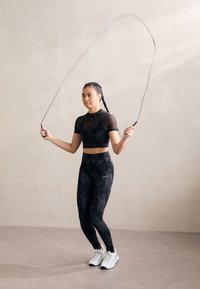 Top corto negro y leggings con un patrón sutil, combinados con zapatillas blancas y una cuerda de saltar en movimiento, todo contra un fondo neutro.