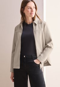 Chaqueta de forro polar gris claro con cuello y cremallera frontal, llevada sobre una blusa negra. Textura lisa. Pantalones vaqueros negros con detalles en los bolsillos.