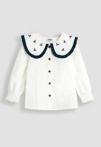 Weißes Langarmshirt mit großem, Rüschensaum und marinegrünem Abschluß sowie bestickten Schleifen. Fünf braune Knöpfe auf der Vorderseite.