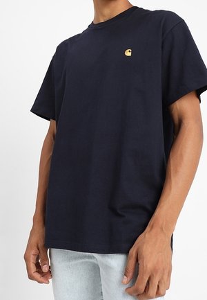 Man wearing a plain dark navy t-shirt with a small yellow logo on the chest, paired with light blue jeans, standing against a white background.