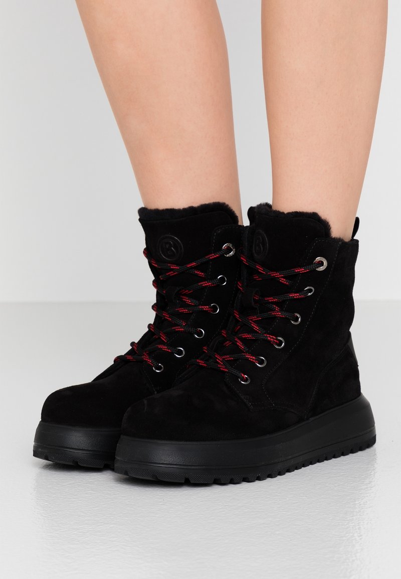 Bottes d'hiver en suède noir avec des lacets rouges et noirs, semelles épaisses et doublure en fourrure, portées sur des jambes nues sur un fond gris clair.