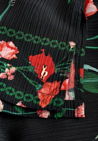 Zwarte stof met een bloemdessin in rood en groen, met een geplooide structuur en een kleine gouden accent met de letter "b".