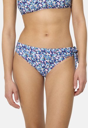 Mujer con braguitas de bikini azul con estampado floral y detalle de lazo lateral, mostrada desde el torso hasta la parte superior de los muslos sobre un fondo sencillo.