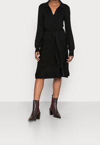 Abito di maglia nera con colletto, maniche lunghe e cintura da annodare. Di lunghezza media con una consistenza liscia. Indossato con stivaletti borgogna alla caviglia.