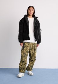 Black puffer jacket with a zippered front and side pockets, paired with loose-fitting green camo pants. White t-shirt underneath, white sneakers.
