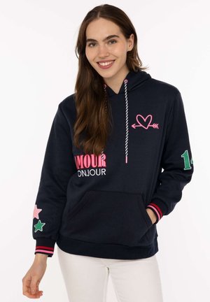 Lächelnde Frau trägt einen marineblauen Hoodie mit pinkem Herz und Pfeil, dem Schriftzug "AMOUR BONJOUR", Stern-Patches auf dem Ärmel und der grünen Nummer 14 am Arm.