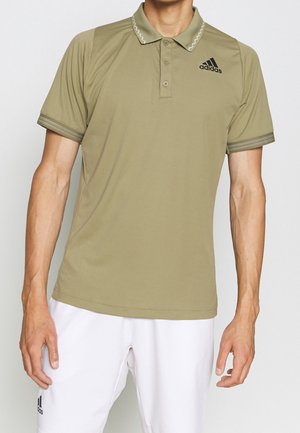 Uomo che indossa una polo Adidas verde oliva e pantaloncini bianchi Adidas, in piedi contro uno sfondo chiaro e uniforme.