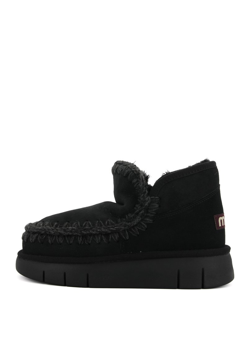 Zwarte suède pantoffel met een bontvoering, decoratieve stiksels en een getextureerde zool. Beschikt over een laag uitgesneden ontwerp en een klein logo aan de zijkant.