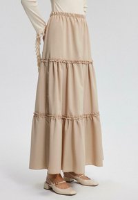 Beige gelaagde rok met Gathered ruffles bij elke naad. De stof is glad, van gemiddelde lengte met een elastische tailleband. Gecombineerd met lichtgekleurde schoenen.