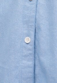 Tissu bleu clair avec une rangée de boutons blancs cousus le long d'une couture verticale, montrant en gros plan la texture et les détails de la couture.