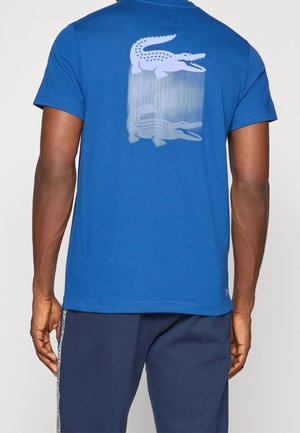 Mand iført blå t-shirt med et lodretstribet hvidt krokodillelogo på øverste del af ryggen og mørkeblå bukser med sidelister.