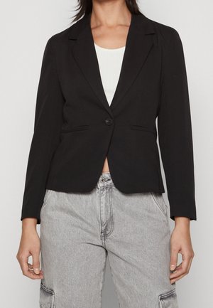Femme portant un blazer noir cintré sur un top blanc et un jean cargo taille haute gris clair, les mains relâchées le long du corps.