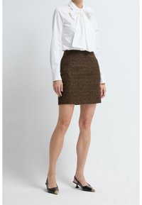Femme portant un chemisier blanc à manches longues avec un grand nœud, une mini-jupe marron texturée et des escarpins slingback noirs avec détail doré.