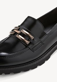 Mocassin en cuir noir avec semelle épaisse et boucle décorative en métal argenté sur le devant.