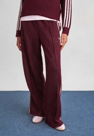 Persona che indossa pantaloni Adidas bordeaux a gamba larga con strisce bianche e logo, abbinati a un maglione coordinato e scarpe rosa su tappeto blu.
