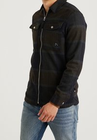 Muž v tmavé kárované bundě na zip s klopovými kapsami a světle modrých džínách, stojící s jednou rukou u pasu.