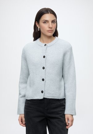 Femme portant un cardigan gris clair boutonné et un pantalon noir, debout devant un fond blanc uni.