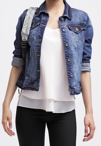 Jasna granatowa kurtka dżinsowa z zapięciem na guziki, dwoma kieszeniami na piersi i podwiniętymi rękawami, założona na białą bluzkę i czarne spodnie.