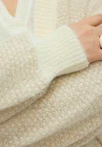 Krēmkrāsas adīts kardigans ar reljefainu rakstu. Audums ir mīksts un pūkains, ar ribotiem piedurkņu manšetēm un biezs, silts izskats.