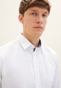Balta ilgomis rankovėmis marška su mygtukų apykakle ir kontrastuojančia tamsia vidine apykakle. Lygi tekstūra ir klasikinis kirpimas.