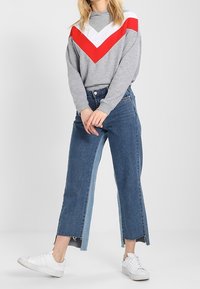 Szara bluza z czerwono-białym wzorem w jodełkę, do tego niebieskie skrócone dżinsy z postrzępionymi brzegami i białe trampki.