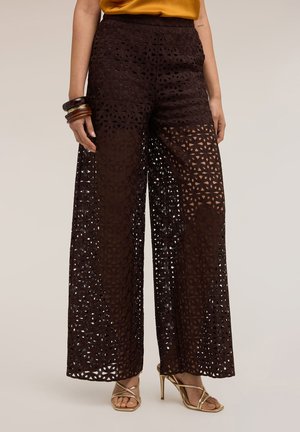 Schwarze Spitzenhose mit hoher Taille und weitem Bein mit geometrischen Ausschnitten, kombiniert mit goldenen Riemchenabsätzen und braunen Armreifen am linken Handgelenk.